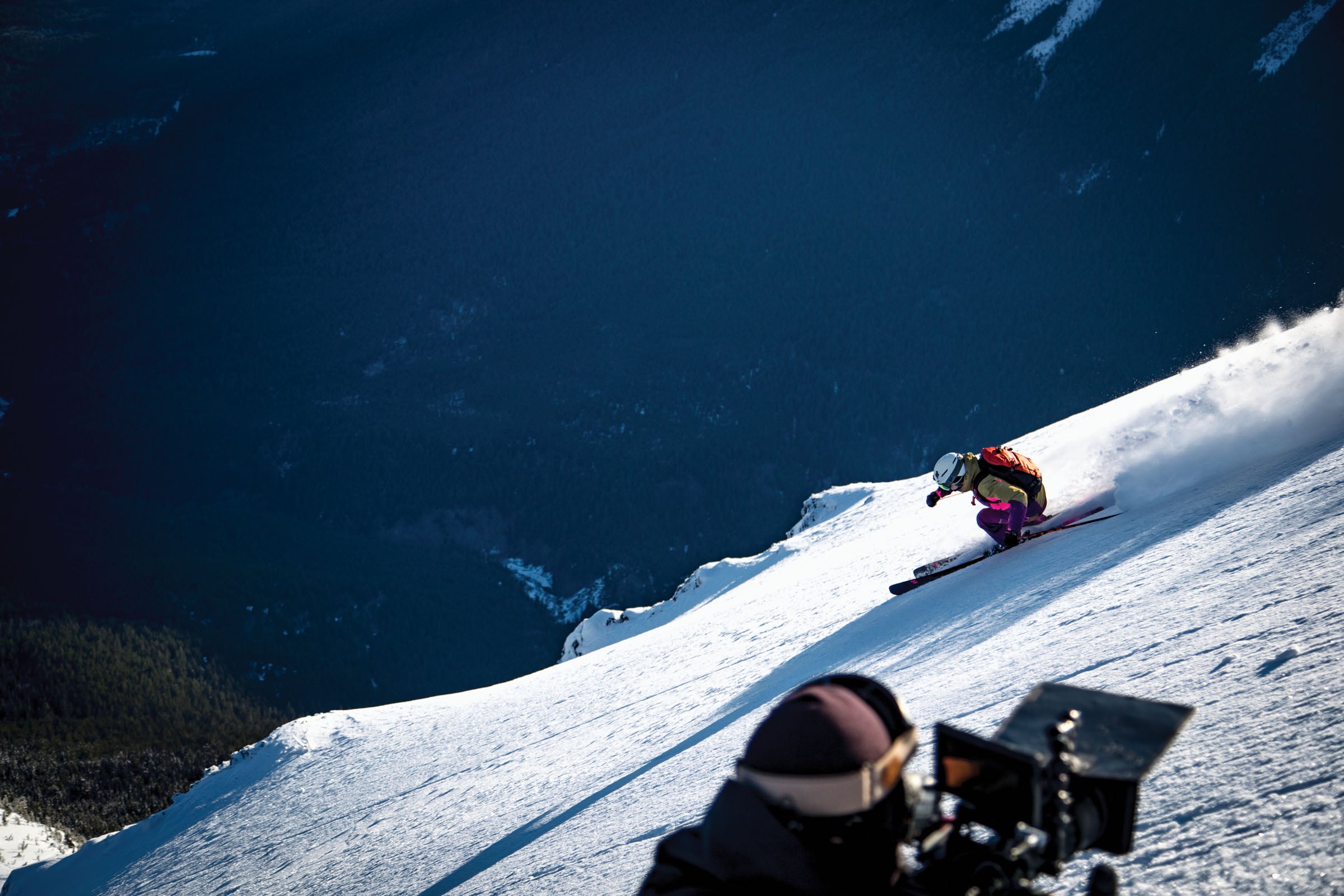 SNW70-BellaCoola-AI0I3195 Amie Engerbretson skiing down slope in front of a videographer