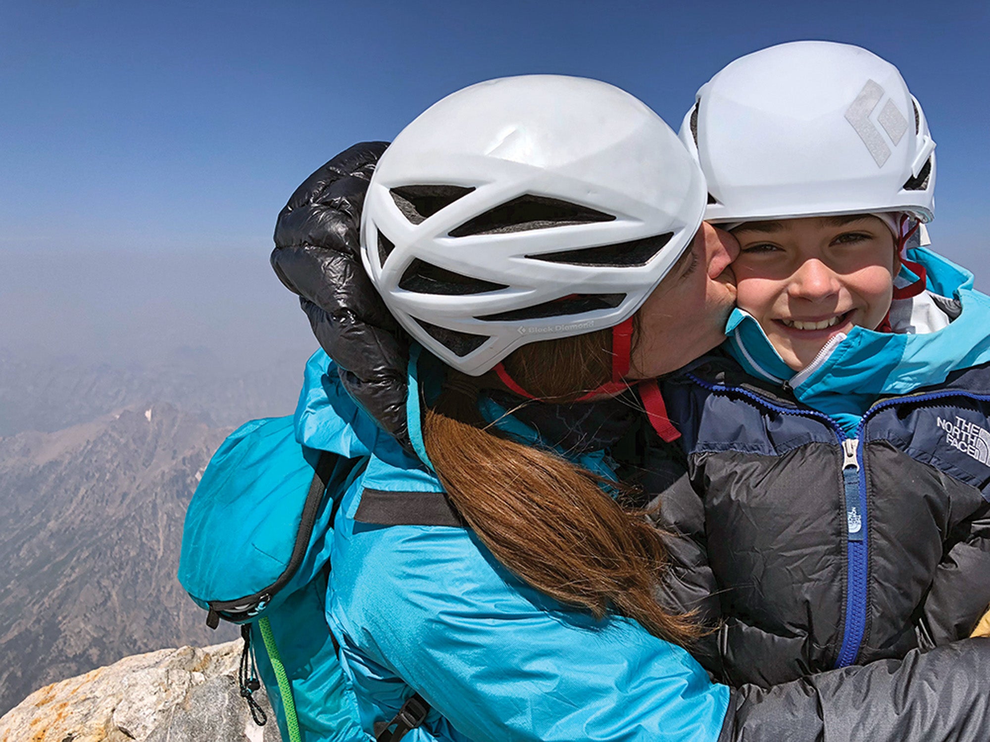 SNW70-Jackson6-coKitDeslauriers copy Kit Deslauriers giving her daughter a huge and a kiss on the cheek in front of a mountain range while they are both wearing helments