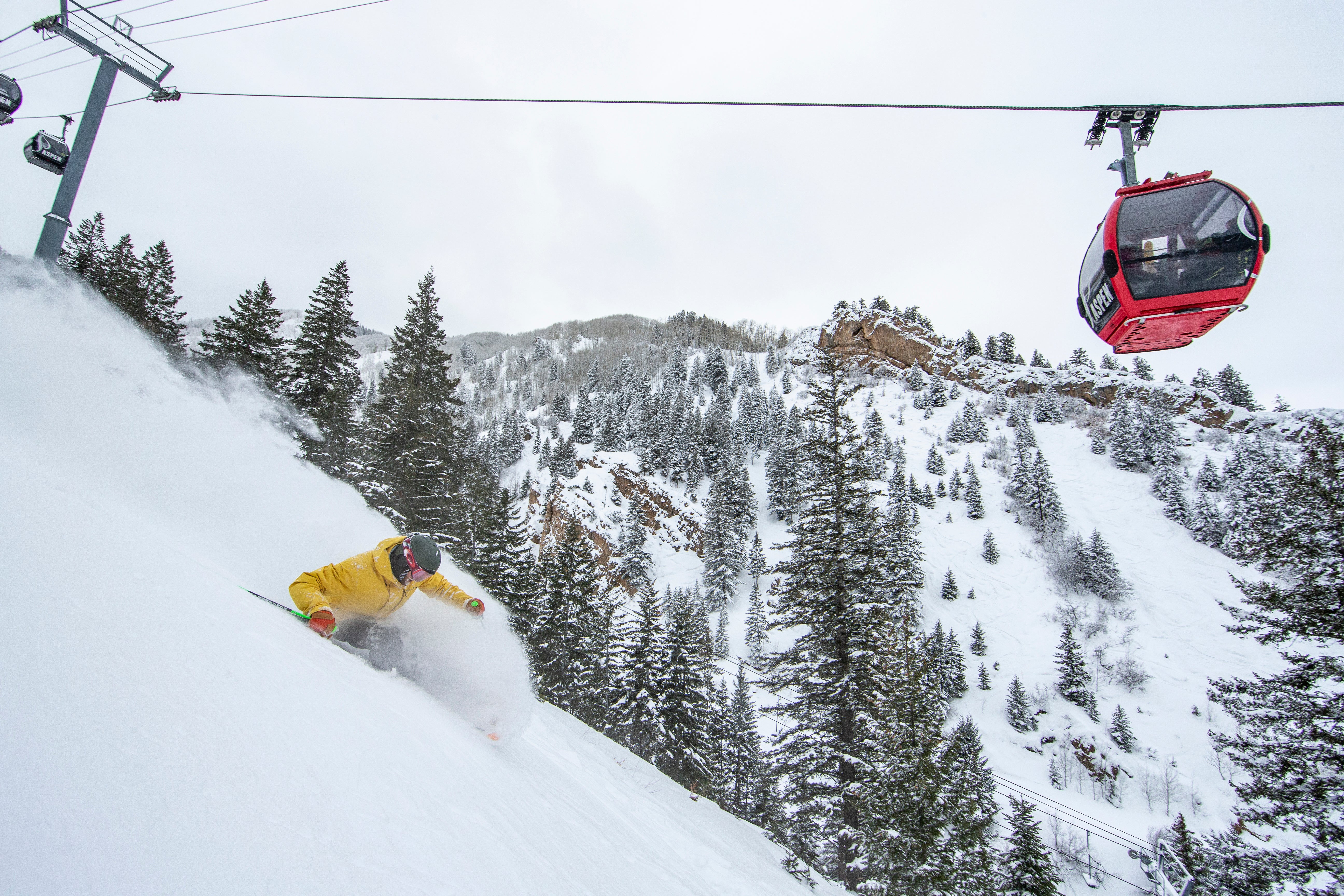 19-A-026 Skier in yellow jacket skiing down slope with a red gondola reading Aspen overhead