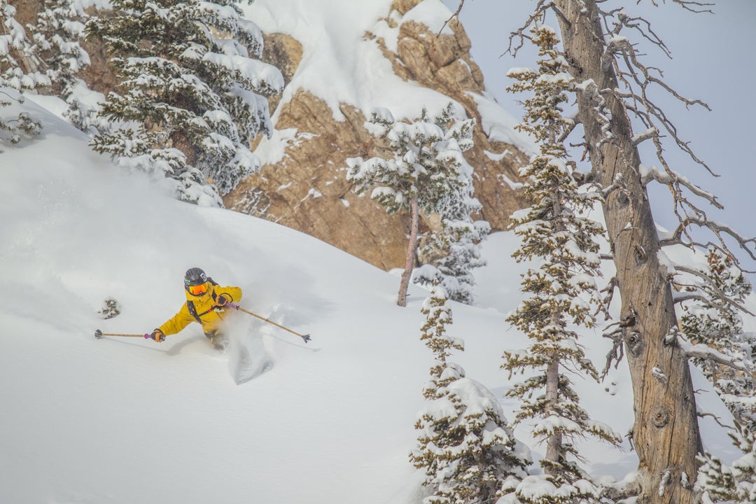 122718-JH-ScottyVermerris-Alta-ALT003 (1 of 1) Person in yellow jacket with orange goggles rips through powder near a rocky cliff and trees at Alta.