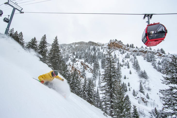 19-A-026 Skier in yellow jacket skiing down slope with a red gondola reading Aspen overhead