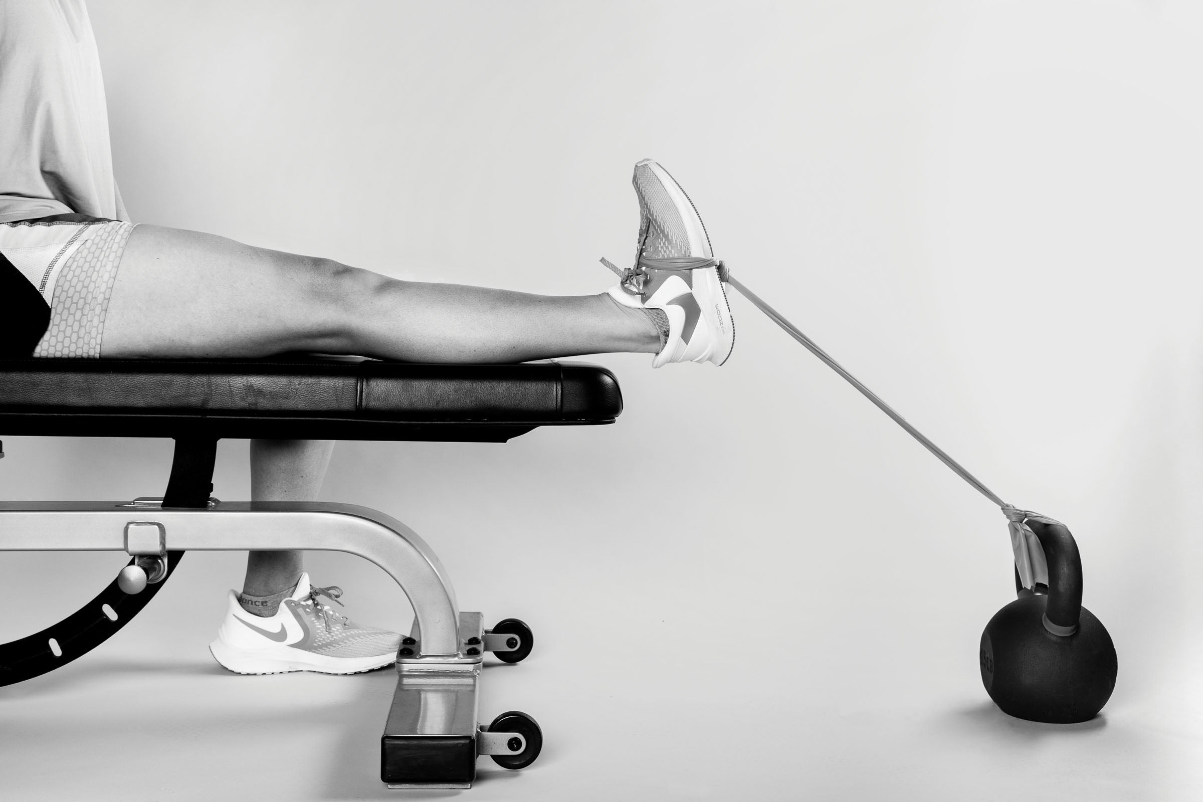 "Black and white photo of someone from the waist down flexing their foot which is tied by a resistance band to a kettle bell."