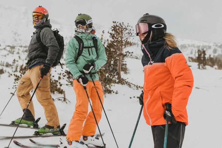 Three people stand on skis at Mammoth Mountain and laugh. All wearing different combinations of black, orange, and light teal. Rolling hills covered…