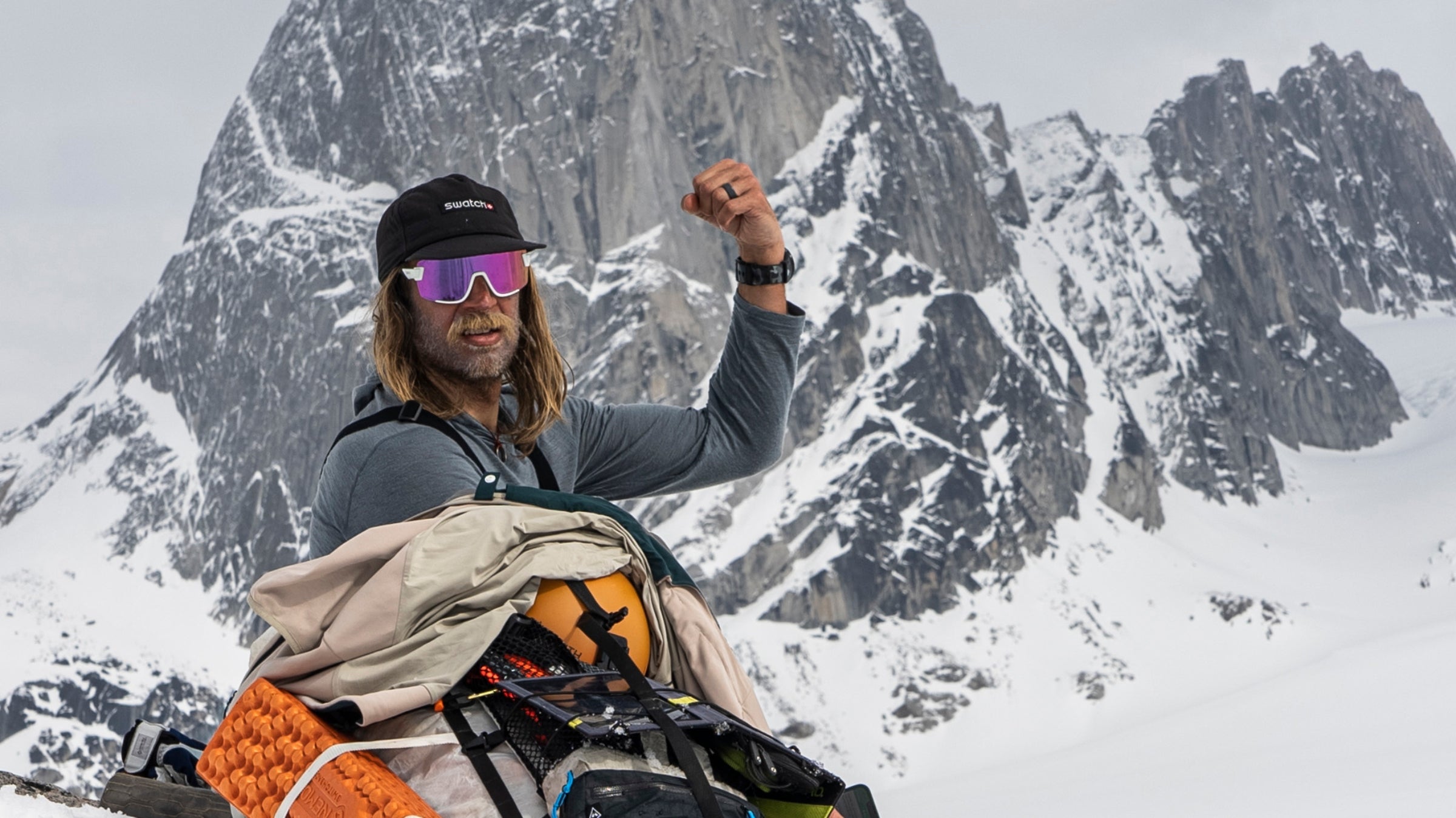 Cody (a white man with long dirty-blond hair, wearing a hat, sunglasses, and ski gear) sits on snow in front of a large, snowy rock face. He flexes his left bicep.