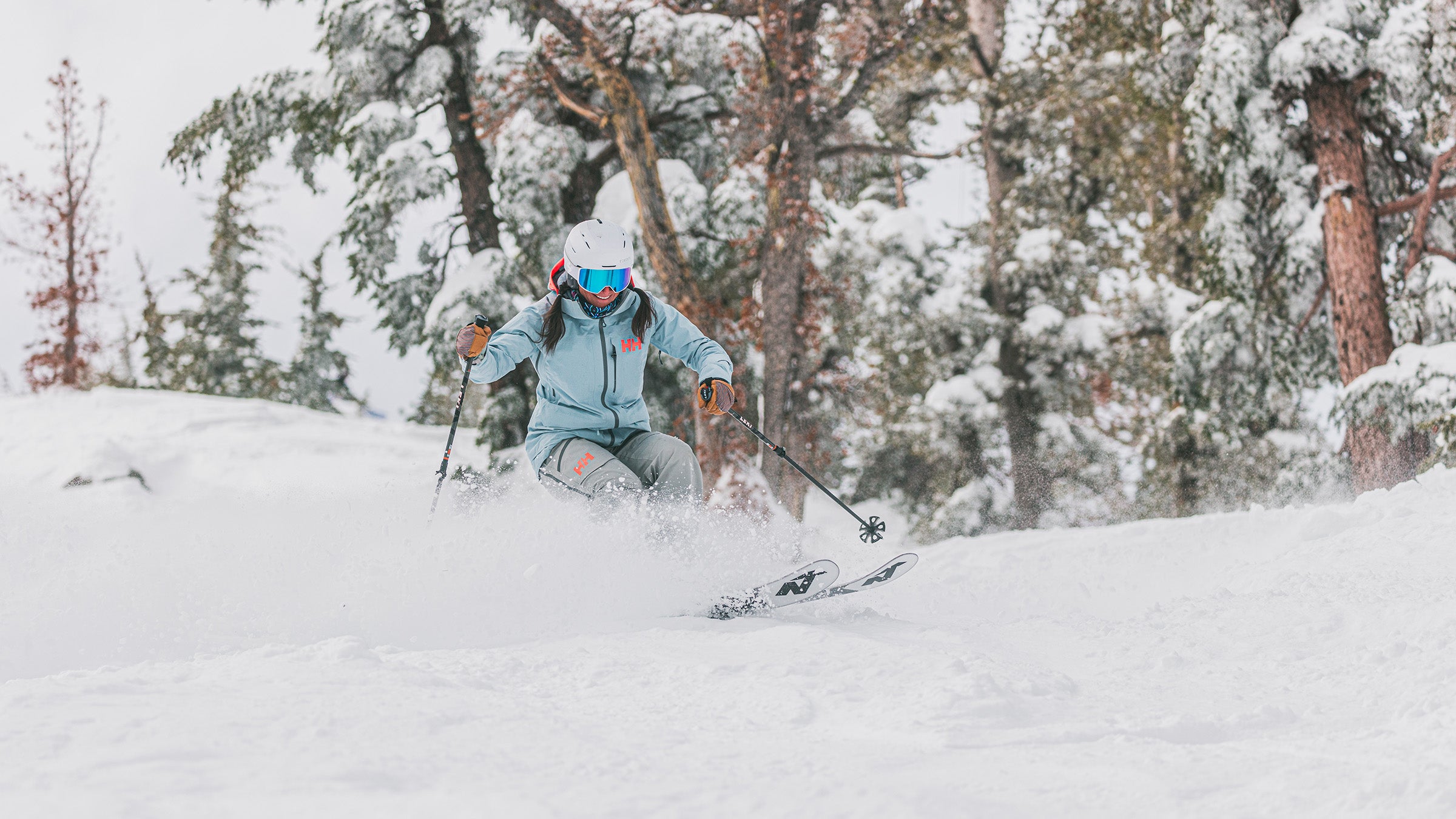 Woman in light blue jacket skiing through powder in trees on wide all-mountain skis